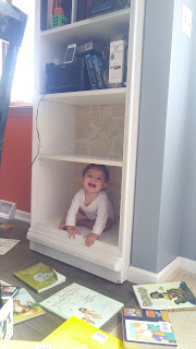 A large bookshelf with the bottom two shelves empty. A number of books are on the floor around it, and sitting on the bottom shelf is a laughing baby.