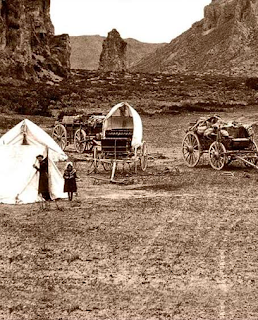 A sepia photograph of a small canvas tent, two small wagons, and curricle. In the background is the entrance to a mountain pass with a small outcropping in the middle like a large pillar. A man wearing a wide-brimmed black hat looks out of the tent opening, and a girl in a bonnet stands in front.