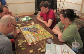 A group of four players sitting around the game board at a a   table, playing Tzolk'in: The Mayan Calendar