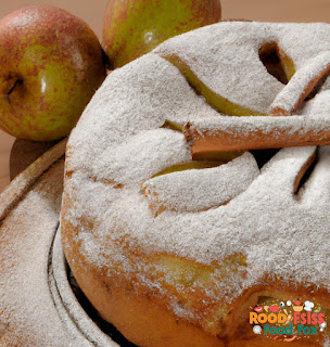 This image shows a freshly baked cinnamon apple cake on a wooden platter, dusted with powdered sugar and garnished with apple wedges and cinnamon sticks.