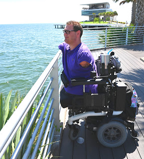 Kyle, in his white-framed, power wheelchair, is wearing a purple Polo shirt. As he looks over the ocean, his right arm is on the railing in front of him. In the background is the St. Petersburg Pier.