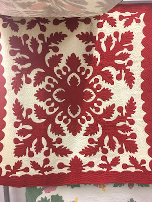 A hanging red and cream Hawaiian applique quilt, with a four-pointed center design of saw-toothed leaves and rounded fruits, and a scalloped, leafy border.