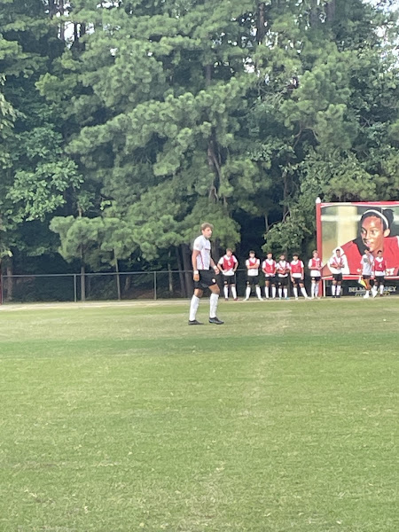 Josh taking a step forward on the field. He does not have the ball. He is wearing his black shorts and white jersey with red lettering.