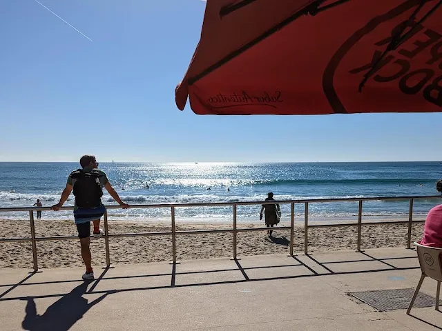 Atlantic Ocean on a sunny day viewed from a restaurant at Carcavelos Beach. There is someone leaning on the railing looking out over the sand in the left of the picture. There is a sun umbrella in the upper right corner of the picture