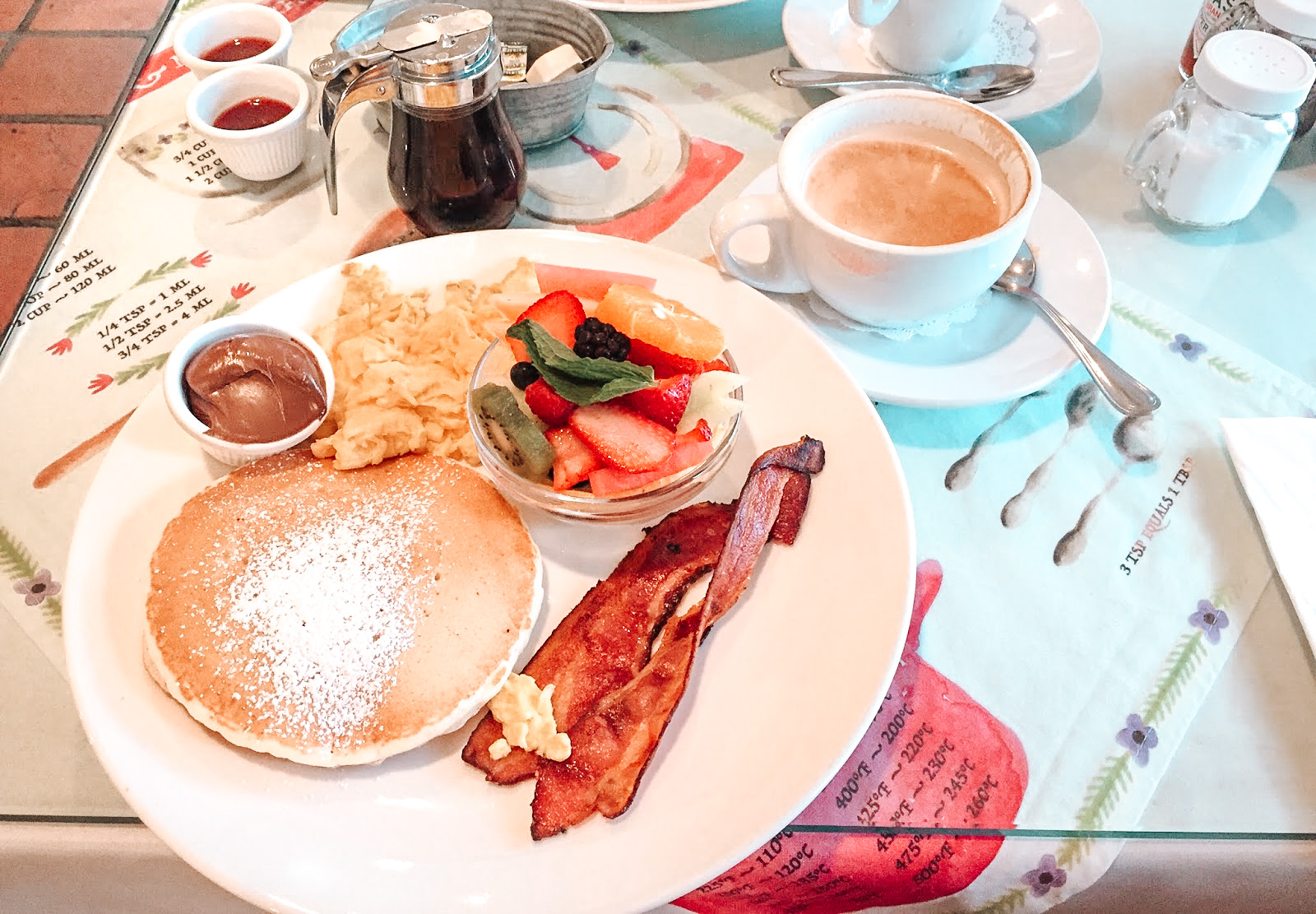 une table du restaurant toast bakery cafe à los angeles avec dans une assiette blanche deux pancakes des oeufs brouillés et du bacon et une coupelle de fruits et en haut à gauche un cappuccino