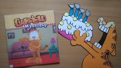 Brązowe tło książka o Garfieldzie i praca plastyczna Garfield trzymający tort  stworzona  na podstawie książek o Garfieldzie