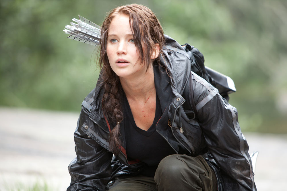 Jennifer Lawrence as Katniss Everdeen crouching down in forest with arrows on her back