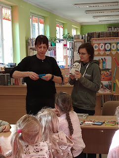Pani przedszkolanka i pani bibliotekarka, pokazują jak wykonać pracę plastyczną. Pani przedzkolanka trzyma w ręku papierową kredkę a pani bibliotekarka pluszową sowę.
