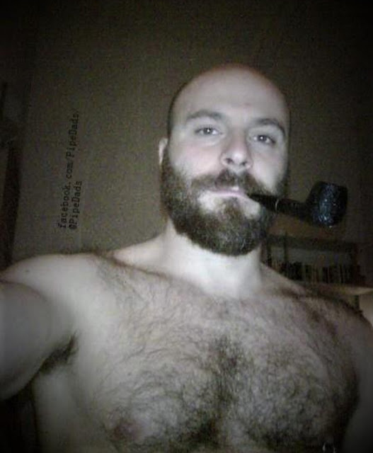 From hairy chest upward a bearded bald daddy with pipe nestled in between teeth taking selfie