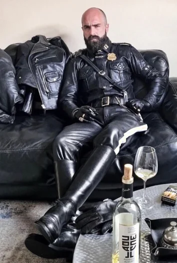 A bearded drunk bald Leatherman sitting on the couch in full gear looking pretty tough