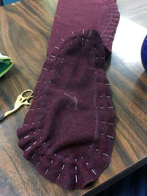 The sole of a garnet-red wool hose, showing densely set pins splayed out around the inch-deep seam allowance like a halo. A pair of gold-handled stork scissors gleam on the brown wood table, and a smudged white chalk R is written upside-down on the center of the sole.