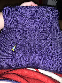 An in-progress hand knit sweater in brilliant purple yarn. The shallow V-neck is centered on an intricate cable panel set between two bias 2 by 2 ribbed sections.