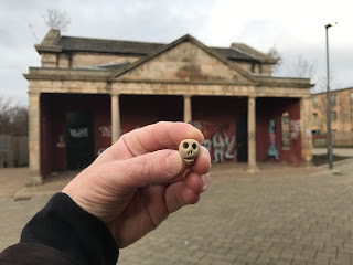 A photo showing a hand holding up a small ceramic skull (Skulferatu 94).  In the background is one of the Leith Fort guardhouses.  Photo by Kevin Nosferatu for the Skulferatu Project.
