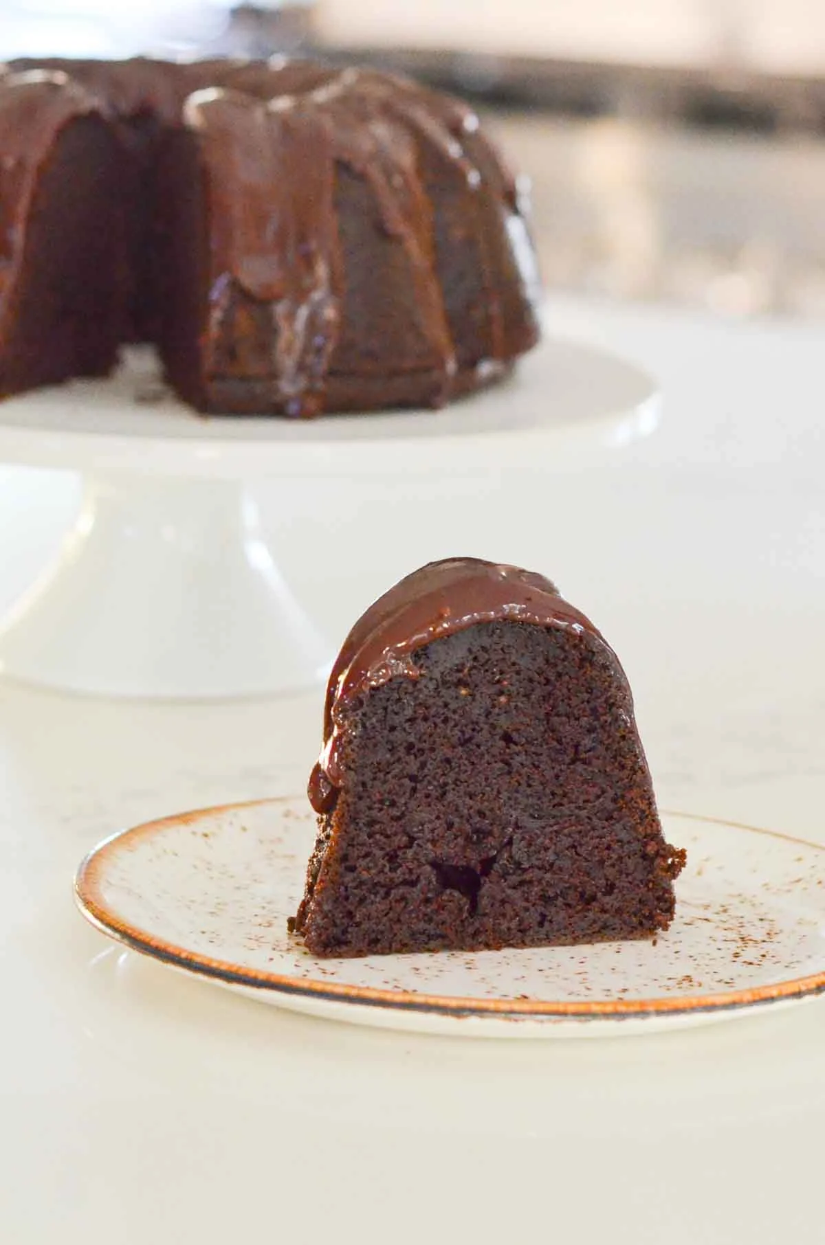 A side shot of a slice of Chocolate Bundt Cake on a plate with the whole cake on a white cake stand in the background.