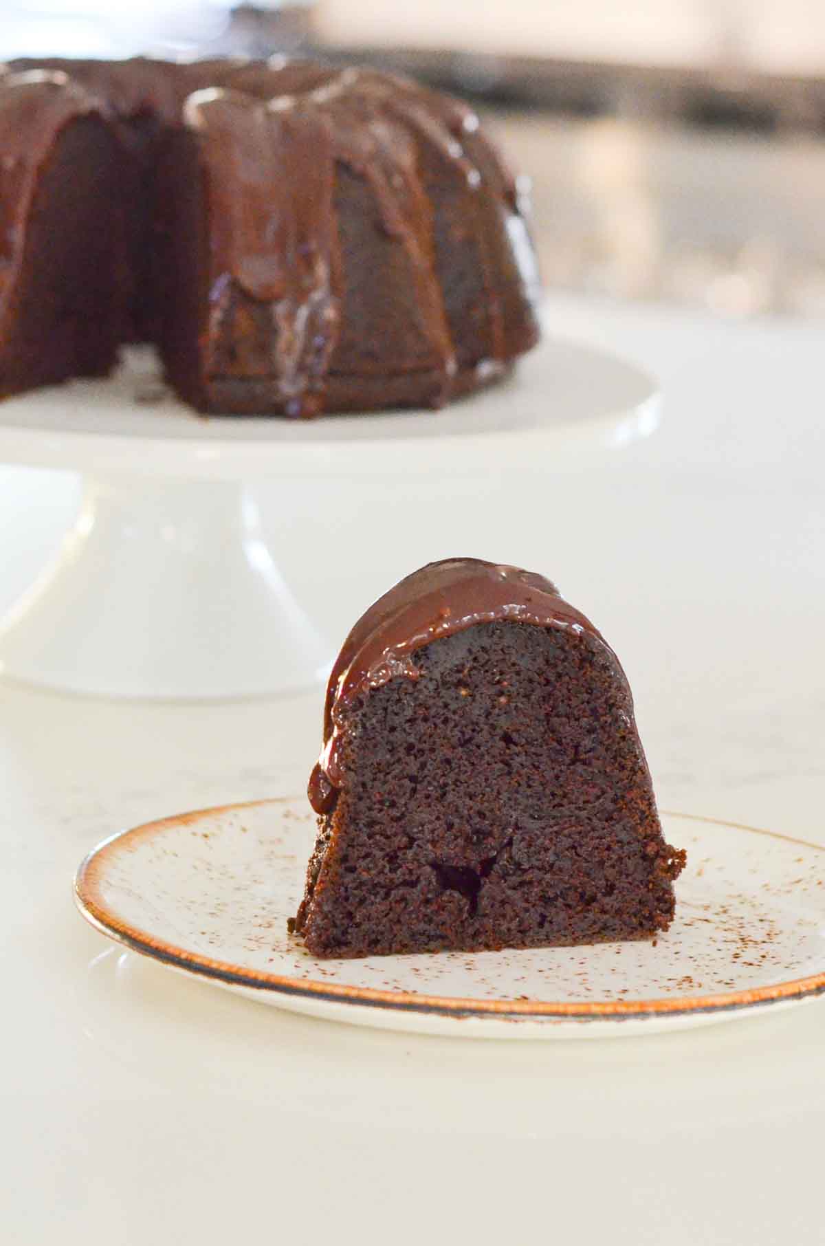 A side shot of a slice of Chocolate Bundt Cake on a plate with the whole cake on a white cake stand in the background.