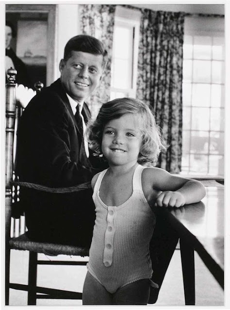 Fotografia em preto e branco do ex-presidente John Kennedy, sentado numa cadeira em frente a uma mesa, e sua filha Caroline ainda criança que está mais próxima da câmera. Ambos sorriem e olham para um ponto à direita deles. O ambiente possui janelas e cortinas, além de um quadro à nossa esquerda.