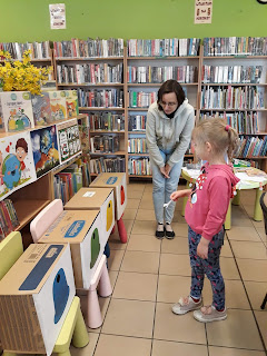 Dziewczynka wrzuca śrubkę do kolorowego pojemnika na odpadki. Tło: Pani bibliotekarka i regały biblioteczne.