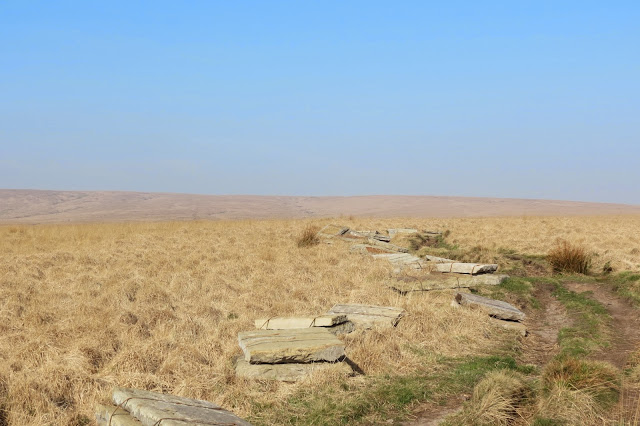 An expanse of moorland as far as the eye can see - pale grass under blue sky. A path of bare earth cuts through the grass and various stones lie at the side of it, ready to be laid. 