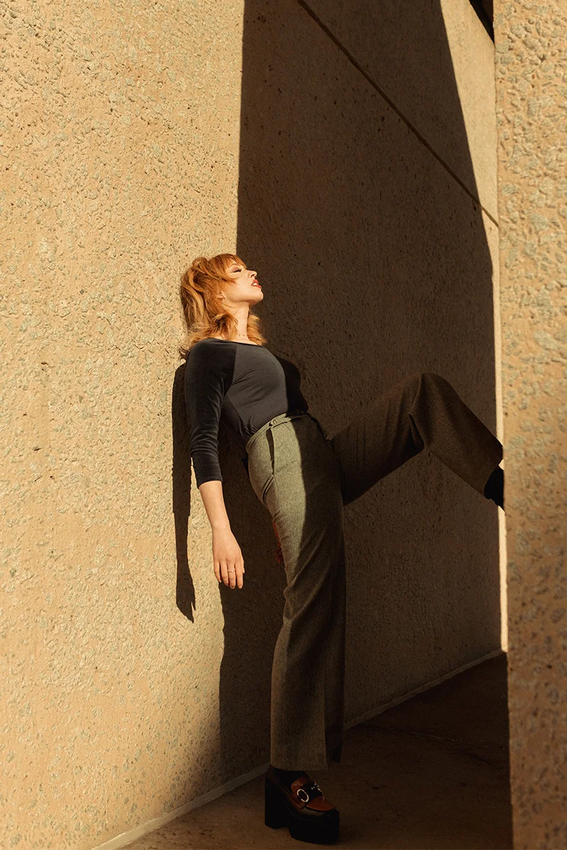 young woman in stylish outfit featuring thin sweater and a pair of high-waist pants posing next to a wall