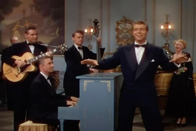 Screenshot from the movie Lullaby of Broadway showing Gene Nelson singing (though his voice was actually dubbed for the movie), standing alongside a blue upright piano and the Page Cavanaugh Trio (piano, guitar, bass). They are all wearing dark suits with bow ties. Nelson has his arms outstretched as he is about to dance as well as sing.