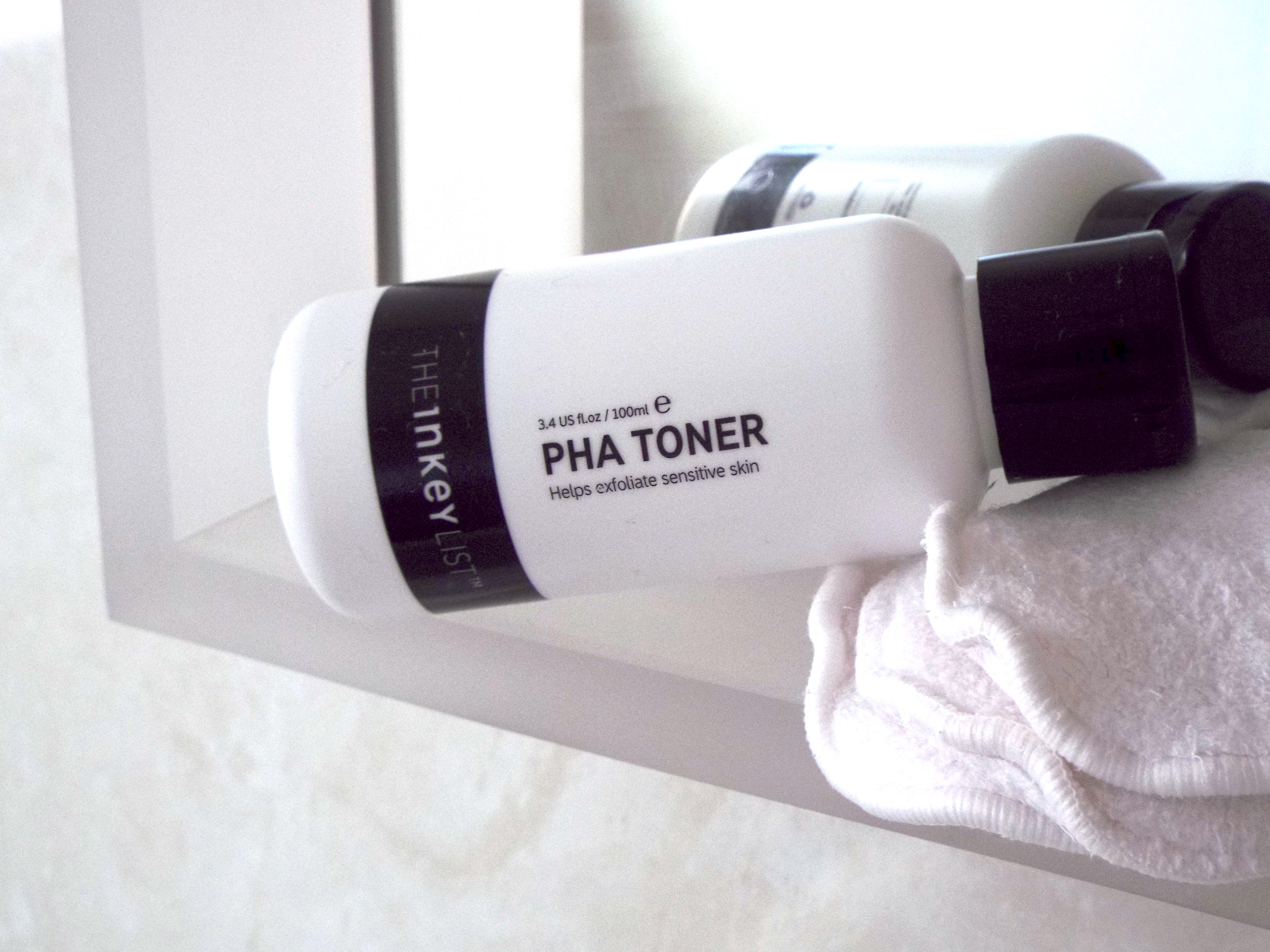 White and black Inkey List PHA Toner bottle lay down on bathroom mirror-side, resting on pile of cotton pads.