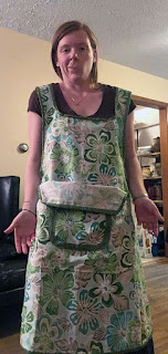 The last of four images in a row, showing a white, redheaded woman at the corner of a hallway, wearing a green, turquoise, and grey bibbed apron bound in light lime-green, with a large near-trapezoidal pocket and a small rounded-square pocket center front, also bound in light lime-green, over a brown t-shirt and long denim skirt. The top edge of the large pocket has flopped down, showing the uneven binding and the back of the fabric, and she's shrugging with palms out toward the viewer, making a 'what can ya do' face..