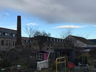 A photo showing a view over the sheds and plants of some allotments, to a red brick chimney with some town houses standing behind it.   Photograph taken by Kevin Nosferatu for the Skulferatu Project.