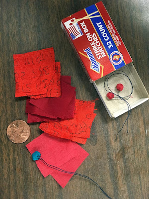 An overhead photo of an open matchbox with two tiny, red-orange fabric buttons in it, a number of red, orange, and pink squares of fabric laid out next to it, a penny that would fit roughly three times on the the fabric squares, and a turquoise fabric button contrasting sharply with the pink fabric under it.