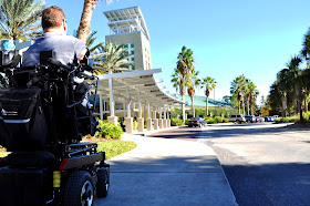 A man in a power wheelchair rolls toward a tall building. Around the building, there are a number of palm trees. The sky is a clear, bright blue.