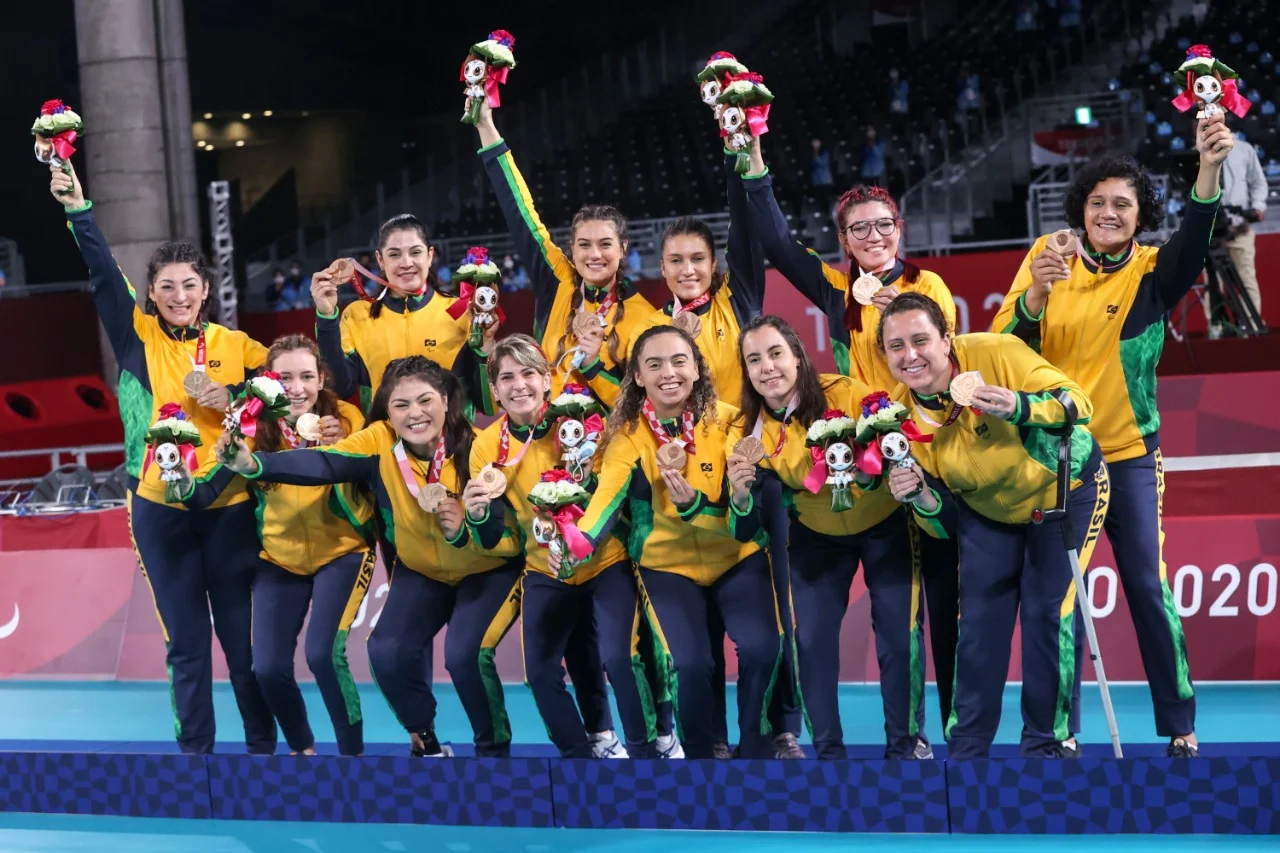 Time de vôlei sentado brasileiro em pé no pódio segurando mascote, medalhas e flores