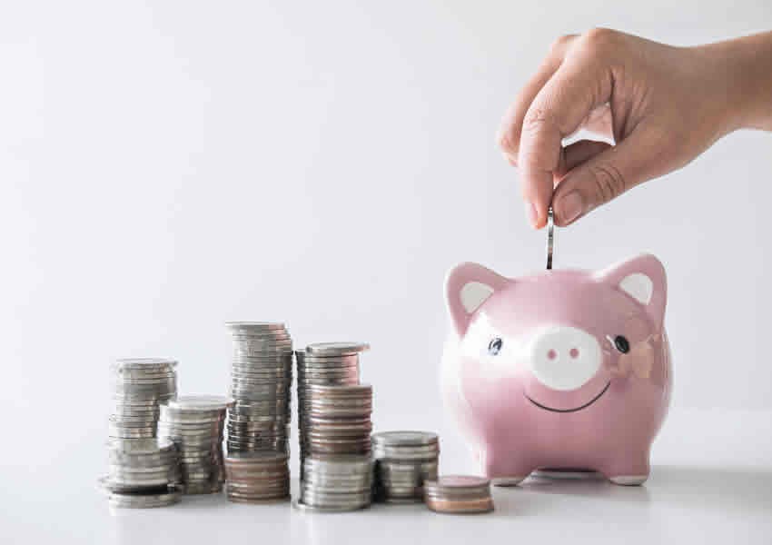 Imagem de cor clara sobre uma mesa uma pilha de moedas ao lado da pilha um cofre porquinho rosa com uma mão sobre ele fazendo o deposito de uma moeda.
