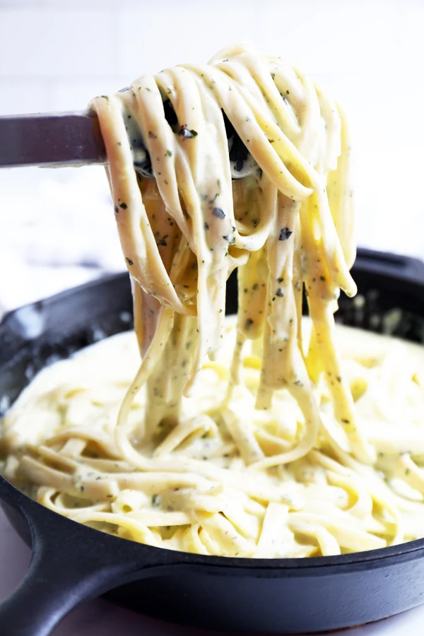 Side view of creamy pesto pasta being lifted with tongs from a sizzling cast iron skillet. The pasta is beautifully coated in a rich and creamy sauce.