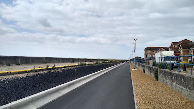 A black asphalt path with a sea wall left and another wall right with houses beyond.