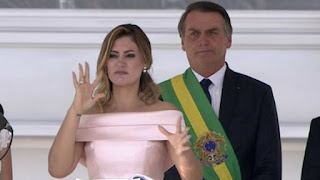 Foto. Ao centro, no palanque e em primeiro plano, Michelle Bolsonaro discursa sinalizando em Libras. Michelle é uma mulher de pele branca, rosto oval, cabelos ondulados castanhos claros com mechas douradas na altura do ombro, sobrancelhas bem delineadas, olhos amendoados castanhos escuros maquiados com sombra preta, nariz levemente arrebitado, lábios médios pintados com batom nude. Usa vestido rosa claro de zibeline de seda com decote de ombro a ombro com detalhe de pala dupla do mesmo tecido, um discreto brinco com pingente de pérola e aliança no anelar direito. Um passo atrás, o presidente empossado de terno escuro sobre camisa branca, gravata e a faixa presidencial usada transversalmente, listrada em: verde, amarelo e verde, ostenta o brasão da República.   Curiosidade: A faixa presidencial foi Instituída em 21 de dezembro de 1910 por um decreto do presidente Hermes da Fonseca, o primeiro a usá-la. E em 21 de Dezembro de 2010, a faixa presidencial brasileira tornou-se centenária e voltou a ter as características originais. Como é confeccionada em seda, o desgaste pelo uso é natural. Desde a época de Collor, a faixa era a mesma e não estava adequada aos moldes definidos no decreto do presidente Hermes. Ao longo dos anos, a faixa presidencial passou por ajustes, ficou mais estreita do que o previsto. Dos 15 cm de largura passou a ter 12,5 cm. O brasão, rico em detalhes e bordado a ouro, ficou simples. Então, o cerimonial sugeriu a confecção de outra peça no centenário, em 2010, nos moldes da faixa original. A faixa foi confeccionada por uma empresa contratada por licitação e custou R$ 38 mil. Ganhou toques de uma restauradora do Instituto do Patrimônio Histórico e Artístico Nacional (Iphan). Ela costurou com fios de ouro o brasão da República na faixa, que é feita em chamalote de seda. As franjas da faixa também possuem pequenas correntes banhadas a ouro. No encontro das extremidades da faixa, um imponente e broche de ouro 18k, maciço, cravejado com 21 brilhantes. No centro, a face da mulher que simboliza a liberdade na pintura de Delacroix “A liberdade guiando o povo. A faixa presidencial deixa seu cofre somente uma vez por ano, no dia 7 de Setembro, quando é usada pelo presidente durante o desfile cívico-militar.