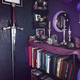 A two-handed claymore sword is mounted on an ornate metal two-pronged mount screwed into a wall clad with black reptile-skin effect wallpaper. The image is looking into a corner. The wall on the right is a dark purple and is stencilled with silver and gold stars. There are three moon-design mirrors in a folk style from Indonesia on the wall, in varying shades of purple with black and gold accents. In the corner there is a three-tier black-painted glossy shelf with various witchy items on it including two resin figures of wizards, several ornate boxes and a statue of Bast. There is a book-case standing from the floor; it is gloss black, and it has a berry purple altar-cloth along the top of it. There is a Neo-Pagan altar set up with a black cast-iron cauldron, an incense censer in the shape of the Moon Goddess, a glass crescent moon candle-holder, several crystals, even more purple glass candle-holders, and a purple incense burner for joss sticks. There are two rolled beeswax candles, purple. The lighting is daylight, the image is relatively bright considering the dark space. Sunlight glints on the faux-reptile wallpaper. There are books on the book-case, disorganised and mis-matched, they are about architecture and include modern books and 19thC volumes
