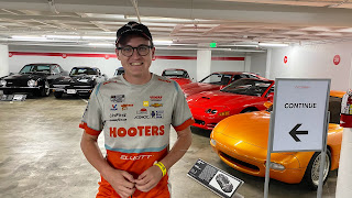 TJ, wearing a racing shirt with the Hooters logo on it, smiles a cheesy smile while surrounded by sports cars in the basement of the auto museum in LA.