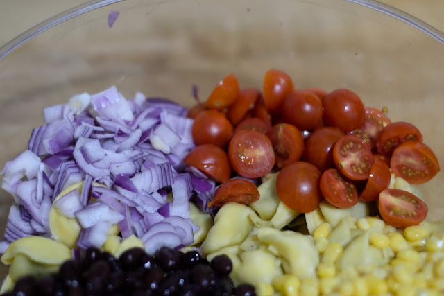 Diced red onion, cherry tomatoes, back beans, corn, and tortellini in a mixing bowl.