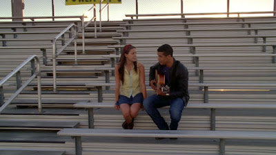 Marley and Jake sitting together in the school bleachers outside as they sing together. Jake is playing the guitar