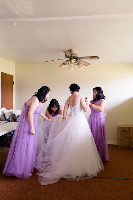 Her bridesmaids are poofing out her dress while she twists to supervise.  The top layers of her dress are a beautiful tulle, giving the illusion that she is floating as she walks.  Her bridesmaids are dressed in lavender soft flowy dresses, each one the same color, but unique in style.  Set at the Southern Oklahoma Chinese Baptist Church in Norman, OK.