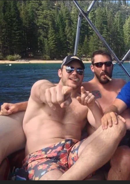 Two shirtless guys sitting on a boat smoking cigars wearing sunglasses