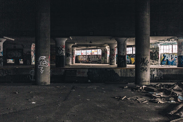 A gloomy view of an abandoned warehouse interior with rows of concrete columns. The space is adorned with vibrant graffiti covering the walls, adding a burst of color to the otherwise monochrome environment. Debris litters the floor, and the absence of active human presence gives the place a haunted atmosphere