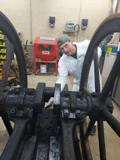 White man wearing white coveralls and green cap is in a garage-type space standing next to a large piece of oil field equipment with a large wheel on each side connected by multiple gears