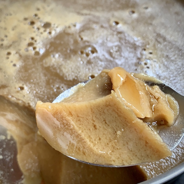Close up of a caramel brown, jelly like dessert being help up with a steel spoon. More dessert in the background. A vegan version of GiNNa (in Kannada), also known as Junnu (in Telegu).