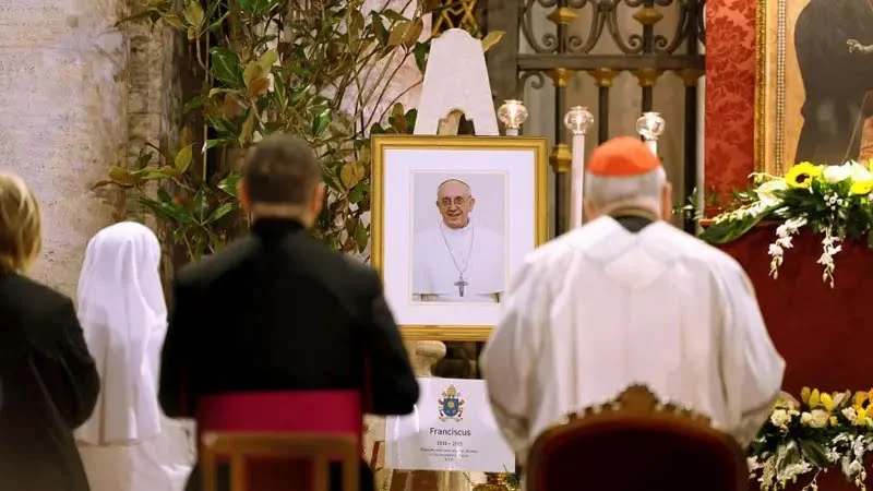Imagem de religiosos em cerimônia com retrato do Papa Francisco ao fundo, destacando o ambiente de oração e reverência.