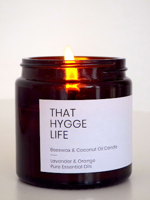Close up shot of the lavender and orange candle, in brown glass bottle, with white paper label, reading That Hygge Life in simple, capital font. The flames burns a golden glow just peaking out of the top of the jar.