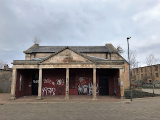 A photo of the frontage of a building, one of the guardhouses.  The front roof is supported by several pillars and the wall is the background is painted red, though id covered in graffiti.  Photo by Kevin Nosferatu for the Skulferatu Project.