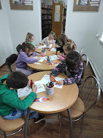 Dzieci w czytelnii Filii Bibliotecznej w Wygiełzowie siedzą przy stolikach i wykonują prace plastyczne - rysunki, zakładki do książek.