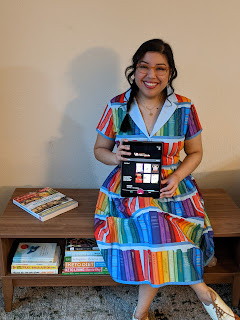 Minerva G., wearing a colorful bookshelf dress, smiling and sitting on a coffee table holding an iPad.