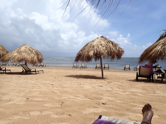 A beach with many palapas and lounge chairs. The only sign of people is my foot at the end of the lounger.