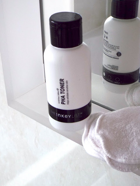 Distant ariel view of black and white Inkey List PHA Toner bottle sat on bathroom mirror-side with cotton pads next to it.